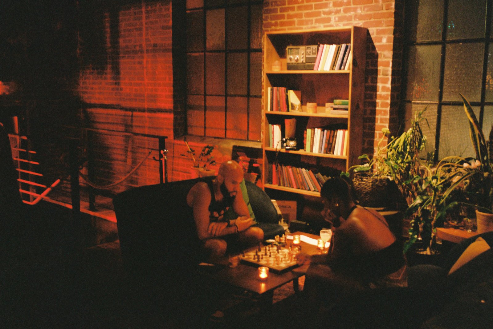 Two guests playing chess by the bookshelf under warm red light